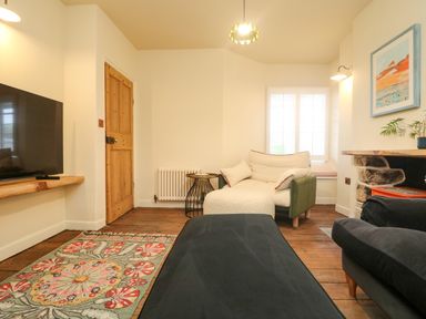 A living room with a patterned rug a wooden door a white cushioned chair a window with shutters a mounted television and a stone fireplace at Josie in Pentewan