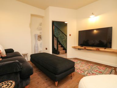 A living room with a black sofa and ottoman a flat screen tv on a wooden shelf and a colorful rug at Josie in Pentewan