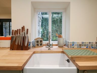 A kitchen sink with a faucet and a window view with a knife block set wooden utensils and a cutting board on a wooden countertop at Josie in Pentewan