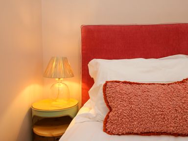 A bedside table with a glass lamp and a bed with white sheets a red headboard and a textured pink cushion at Josie in Pentewan