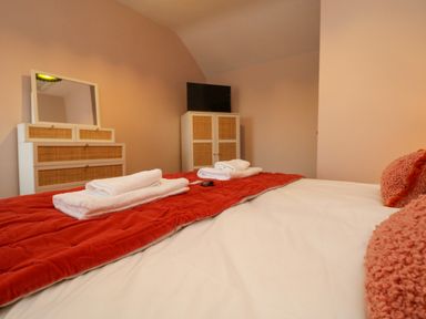 A bedroom with a bed with white sheets and red pillows and a red blanket with towels on top a dresser with a mirror and a cabinet with a television at Josie in Pentewan