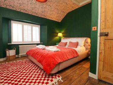 A bedroom with a bed with red bedding and pillows a patterned rug a wooden door and a window with shutters at Josie in Pentewan