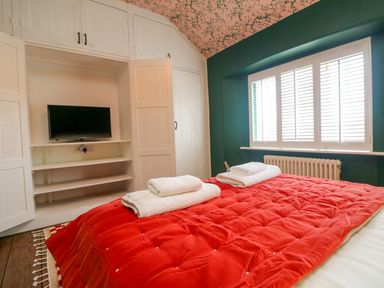 A bedroom with a red bedspread white towels a built in wardrobe with a television and shuttered windows at Josie in Pentewan