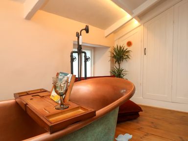 A copper bathtub with a wooden bath tray holding a glass and book in a bathroom with wooden floor and built-in cupboards at Josie in Pentewan