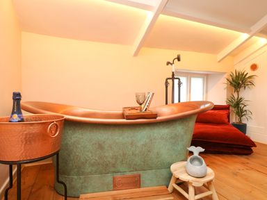 A bathroom with a copper bathtub a copper bucket with a bottle a wooden stool with a whale shaped soap holder a red cushion and a plant at Josie in Pentewan