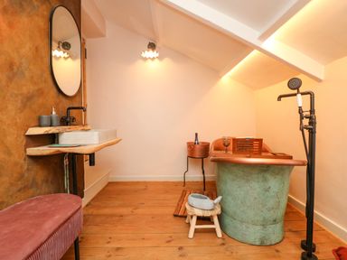 A bathroom with a freestanding bathtub a wooden stool a round mirror a wall-mounted sink and a bench at Josie in Pentewan