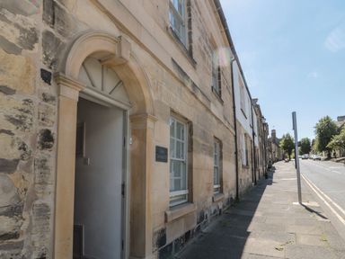 Stone building with arched doorway and windows along a sidewalk on a street with trees and cars at Eider Cottage in Warkworth