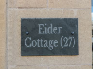 A black sign with white lettering reading Eider Cottage 27 mounted on a stone wall at Eider Cottage in Warkworth