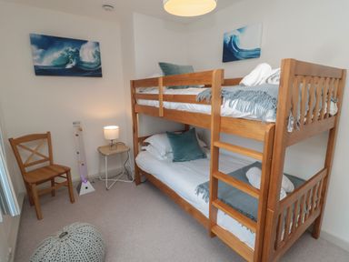 A bedroom with wooden bunk beds a wooden chair a small side table with a lamp a knitted pouf and wave paintings on the walls at Eider Cottage in Warkworth