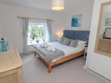 A bedroom with a double bed with a blue headboard and gray pillows next to a window with curtains and a wooden dresser at Eider Cottage in Warkworth