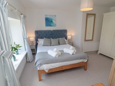 A bedroom with a double bed with a blue headboard and three pillows with bedside tables and lamps and a window with striped curtains at Eider Cottage in Warkworth
