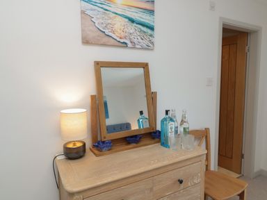 A wooden dresser with a mirror a lamp bottles and glasses next to a wooden chair and a doorway at Eider Cottage in Warkworth