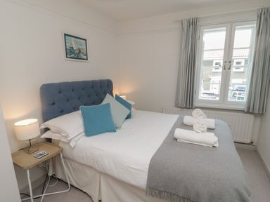 A bedroom with a double bed blue headboard pillows rolled towels bedside table lamp and window with curtains at Eider Cottage in Warkworth