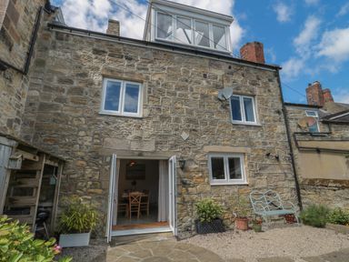 A stone exterior of a house with open double doors leading to a dining area and a bench with plants in pots outside at Eider Cottage in Warkworth