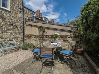 An outdoor patio with a round table and six chairs with blue cushions next to a stone wall at Eider Cottage in Warkworth