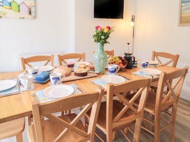 A dining table set with plates cups bread and fruit in a room with wooden chairs at Eider Cottage in Warkworth