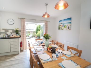A dining room with a wooden table set for six next to kitchen cabinets and a door opening to a garden at Eider Cottage in Warkworth