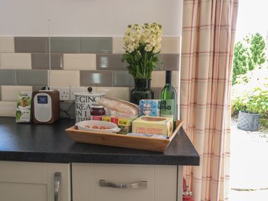 A kitchen countertop with a wooden tray holding food items and a green vase with flowers next to a radio and tiled wall with outlets at Eider Cottage in Warkworth