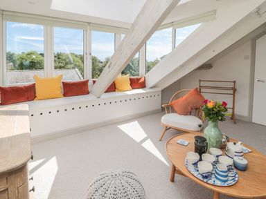A cozy sitting area with a wooden chair and colorful cushions by large windows and a wooden table with tea set and flowers at Eider Cottage in Warkworth