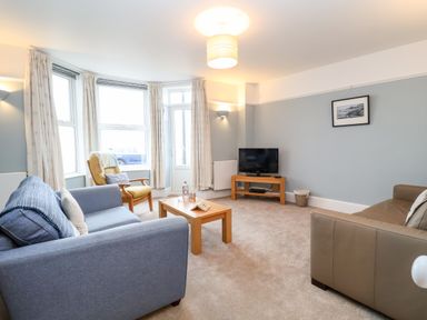 A living room with a blue sofa, a brown chair, a small wooden table with a bottle and glasses, a television on a wooden stand, and framed picture on the wall at 11 The Craighurst in Southwold