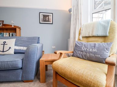A living room with a blue sofa with anchor cushion and a yellow armchair with a pillow by the window at 11 The Craighurst in Southwold