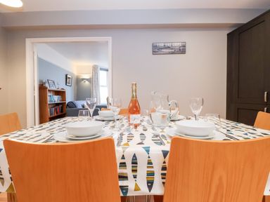 A dining table set with plates glassware and a bottle with wooden chairs in a dining room with a view into a living room at 11 The Craighurst in Southwold