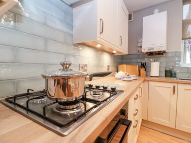 A kitchen with a gas stove and a pot on it white cabinets a knife block a teapot with cups and a boiler on the wall at 11 The Craighurst in Southwold