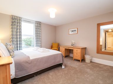 A bedroom with a bed, wooden desk, armchair, window with patterned curtains and a large mirror at 11 The Craighurst in Southwold