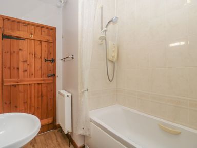 A bathroom with a bathtub and shower, a white sink, a wooden door, and a radiator at Anvil Cottage in Gatcombe near Blakeney