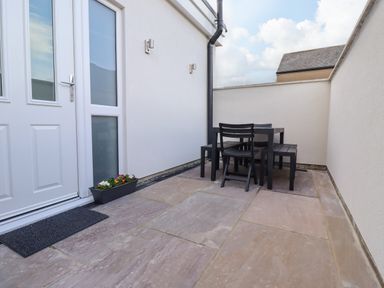 An outdoor patio with stone flooring a black dining table with chairs and benches next to a white door with a mat and a flower pot at 2A Front Street in Embleton