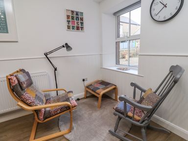 A seating area with a wooden chair, gray rocking chair, footstool, floor lamp, wall clock, and window at 2A Front Street Embleton
