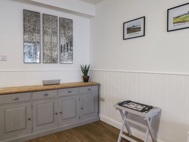 A room with a grey sideboard with drawers and cabinets a small plant on top three vertical mirrors on the wall and two framed pictures above a folding tray table at 2A Front Street in Embleton