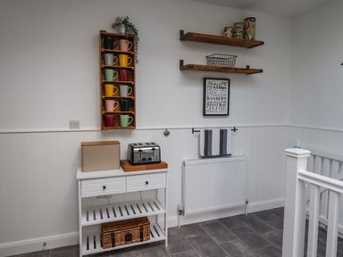 A kitchen area with a white cabinet holding a toaster and bread box with mugs on wall shelves and a towel above a radiator at 2A Front Street in Embleton