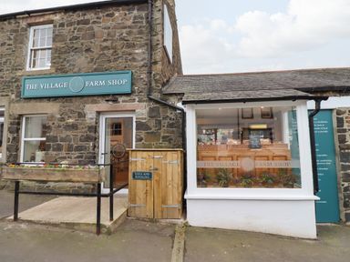 Stone building with a sign reading The Village Farm Shop and a wooden planter with flowers at 2A Front Street in Embleton