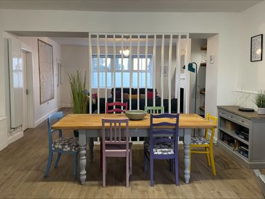 A dining area with a wooden table and colorful chairs facing a living room with sofas separated by vertical white slats at 2A Front Street in Embleton