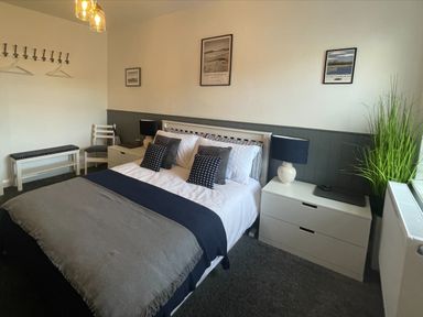 A bedroom with a bed pillows side tables lamps a chair a bench and framed pictures on the wall at 2A Front Street in Embleton