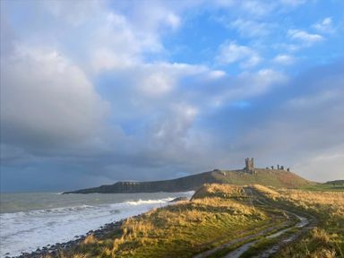 A coastal landscape with a hill, ruins on top, a dirt path, grass, and the sea under a cloudy sky at 2A Front Street in Embleton