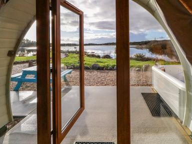 An open glass door looking out to a patio with a blue picnic table and a hot tub beside a lake at The Wheelhouse Pod No 1 in Burtonport County Donegal
