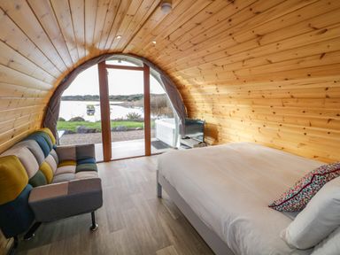 An arched wooden room with a bed, sofa, small TV, and glass door overlooking a lake at The Wheelhouse Pod No 1 Burtonport County Donegal