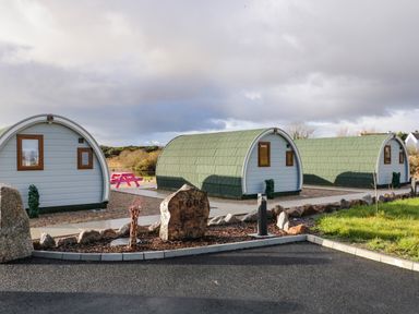 Three pod-shaped cabins with green roofs and white walls near a stone sign that says pods at The Wheelhouse Pod No 1 in Burtonport County Donegal