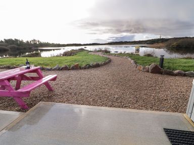 A gravel path with stone edging leading to a body of water with a small boat and a pink picnic table on a grassy area at The Wheelhouse Pod No 2 Burtonport County Donegal