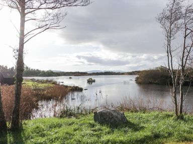 A lakeside scene with grass trees a boat on water and distant hills at The Wheelhouse Pod No 2 Burtonport County Donegal
