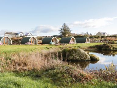 An outdoor scene with small pod-shaped cabins and picnic tables near a pond at The Wheelhouse Pod No 3 in Burtonport County Donegal