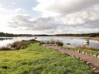 A grassy area with a gravel path and bench leading to a lake with a small boat at The Wheelhouse Pod No 3 Burtonport County Donegal