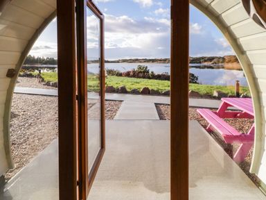 A view through open wooden doors showing a gravel path green grass and a body of water with a pink picnic table outside at The Wheelhouse Pod No 7 Burtonport County Donegal
