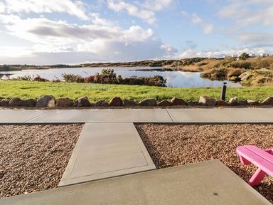 A paved patio area with gravel and a pink bench overlooking a lake and grassy land at The Wheelhouse Pod No 7 Burtonport County Donegal