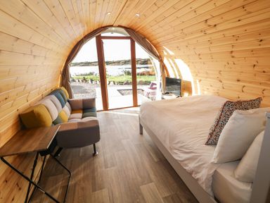 A wooden pod interior with a bed and a multicolored sofa near glass doors opening to a waterside view at The Wheelhouse Pod No 9 in Burtonport County Donegal
