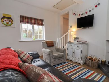 A living room with a leather sofa red throw cushions armchair striped rug small cabinet and wall mounted television at The Little White Cottage in Milton-Under-Wychwood