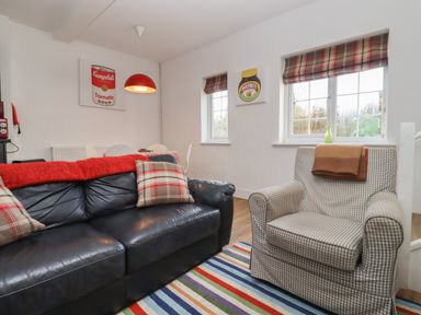 A living room with a black leather sofa and checked armchair near windows with plaid blinds at The Little White Cottage in Milton-Under-Wychwood