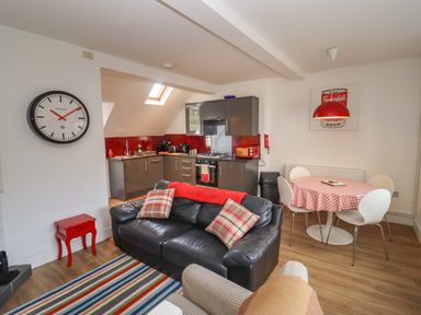 An open plan living room with a black sofa and striped rug beside a kitchen with grey cabinets and red backsplash at The Little White Cottage in Milton-Under-Wychwood